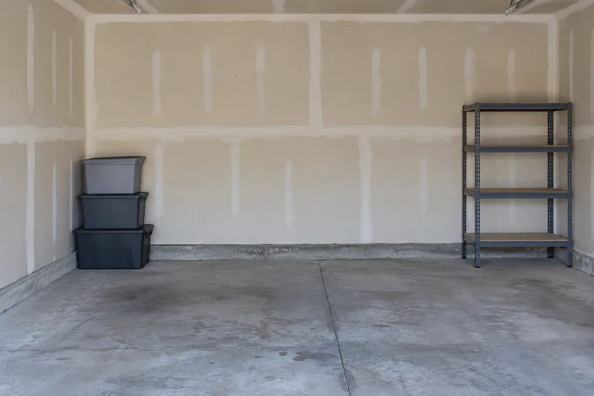 Bare garage wall of taped drywall with plastic storage bins stacked on the concrete floor and a metal shelving unit